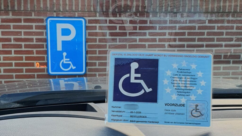 Afbeelding van een gehandicaptenparkeerkaart achter het raam van een auto die geparkeerd staat voor een blauw bordje met daarin de letter P en een rolstoelsymbool wat aanduidt dat je daar mag parkeren met een gehandicaptenparkeerkaart.