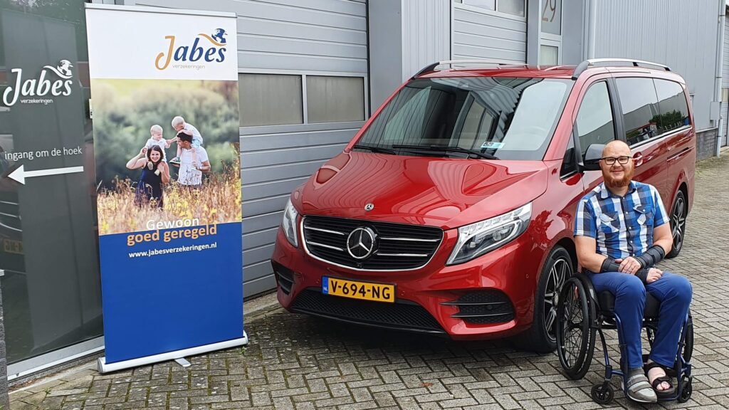 afbeelding van Ralph Stoové in zijn rolstoel zittend naast zijn rode Mercedes-Benz V-Klasse rolstoelbus en een banner van tussenpersoon Jabes Verzekeringen