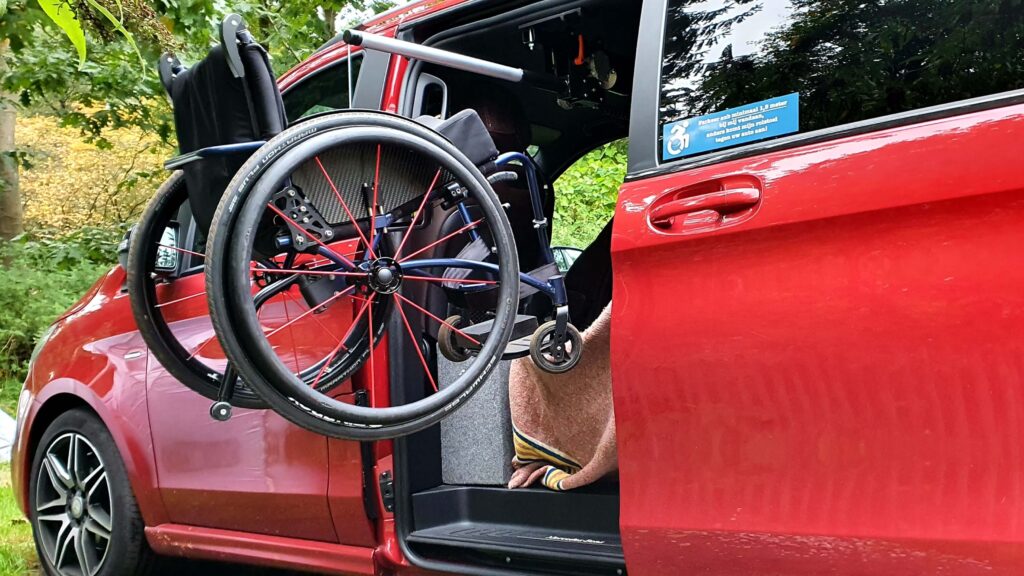 Afbeelding van een rolstoel met rode spaken in Spinergy rolstoelwielen hangend in een rolstoelliftje van een rolstoelbus die een rolstoelsticker op de zijruit heeft met de aanduiding om afstand te houden ivm in- en uitladen van de rolstoel.