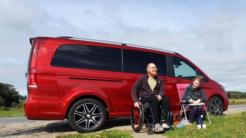 Afbeelding van een man in een rolstoel met rode Spinergy wielen en een vrouw die voor een hyasintrode Mercedes-Benz V-Klasse rolstoelbus zitten