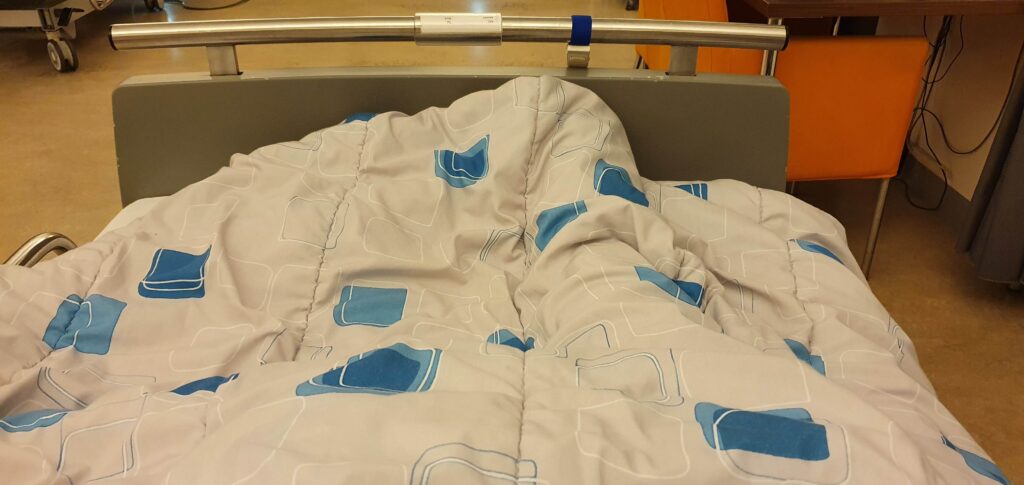 afbeelding van patiënt in een ziekenhuisbed waarvan onder de dekens net zichtbaar is dat het linker onderbeen is geamputeerd