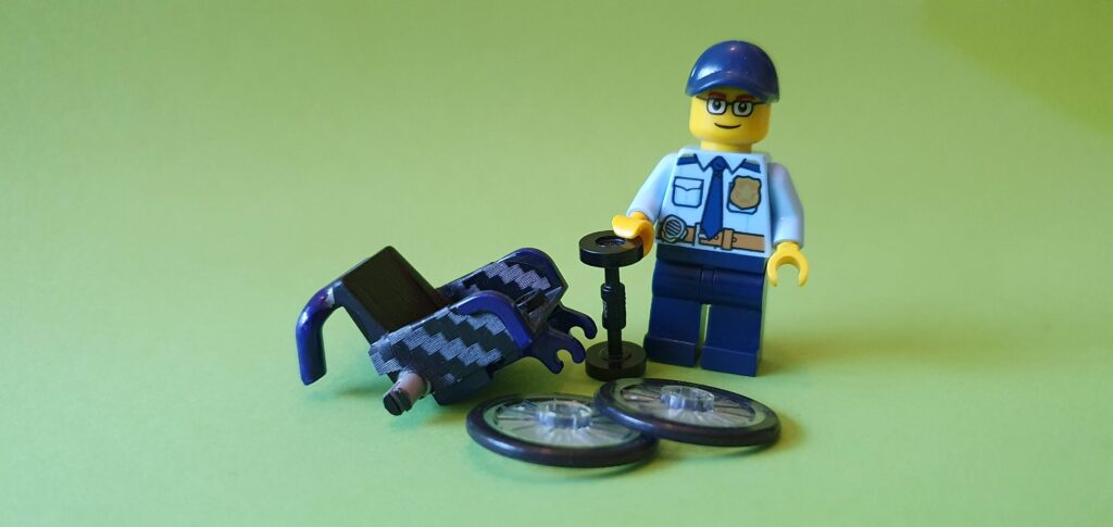 afbeelding van LEGO SigFig staande bij een rolstoeltje in onderdelen, maar al wel geverfd in blauw en zwart zoals de echte rolstoel van Ralph Stoove