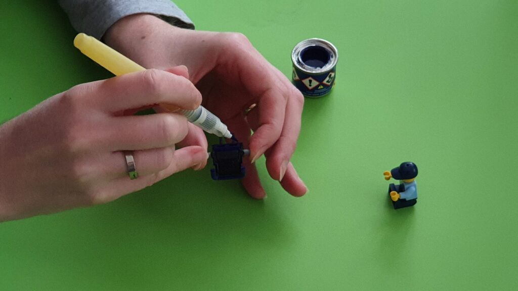 Afbeelding van twee handen die met een penseel en modelbouw verf van Revell een grijs LEGO rolstoeltje in parelmoer nacht blauw en zwart verven, zodat die lijkt op de echte rolstoel van RaGaSto tbv de SigFig in politie uniform rechts op de foto.