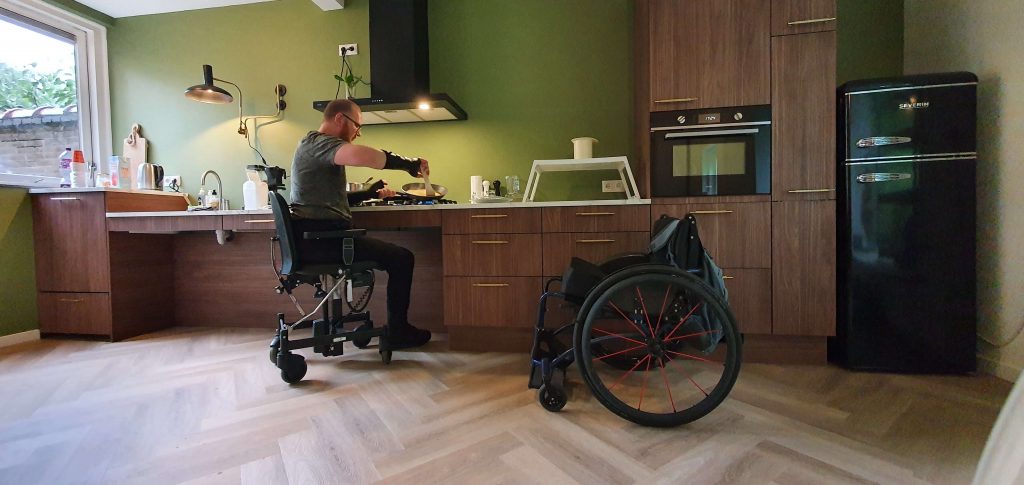 foto van een man die in een trippelstoel kookt bij een houtkleurige aangepaste en onderrijdbare keuken met daarnaast zijn lege rolstoel tbv disability pride month
