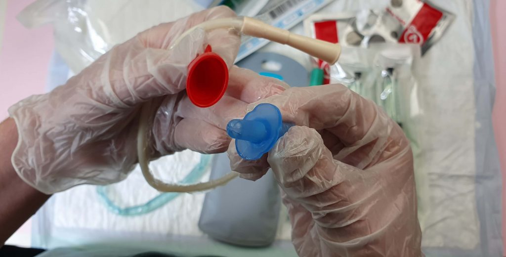 Een close-up van een eenmalige katheter vastgehouden door twee handen in steriele handschoenen met op de ondergrond diverse verpakkingen van verschillende merken en soorten katheters
