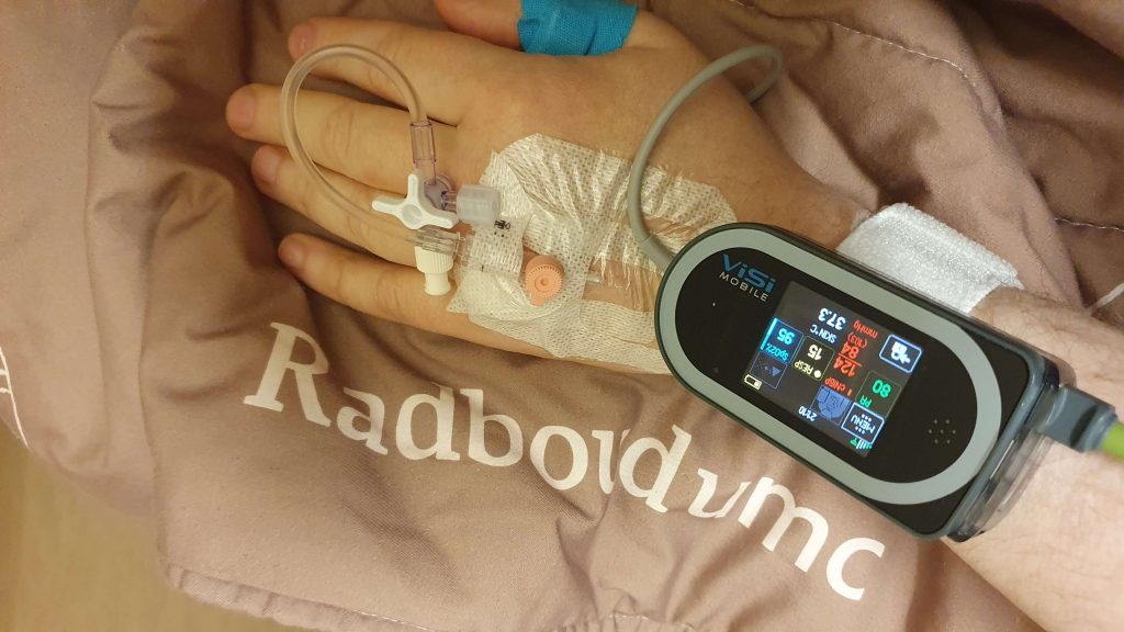 afbeelding van een hand met een infuus met een modern meetkastje dat de vitale functies meet zoals bloeddruk, hartslag, saturatie en lichaamstemperatuur. De hand ligt op een dekbed van het ziekenhuis RadboudUMC.