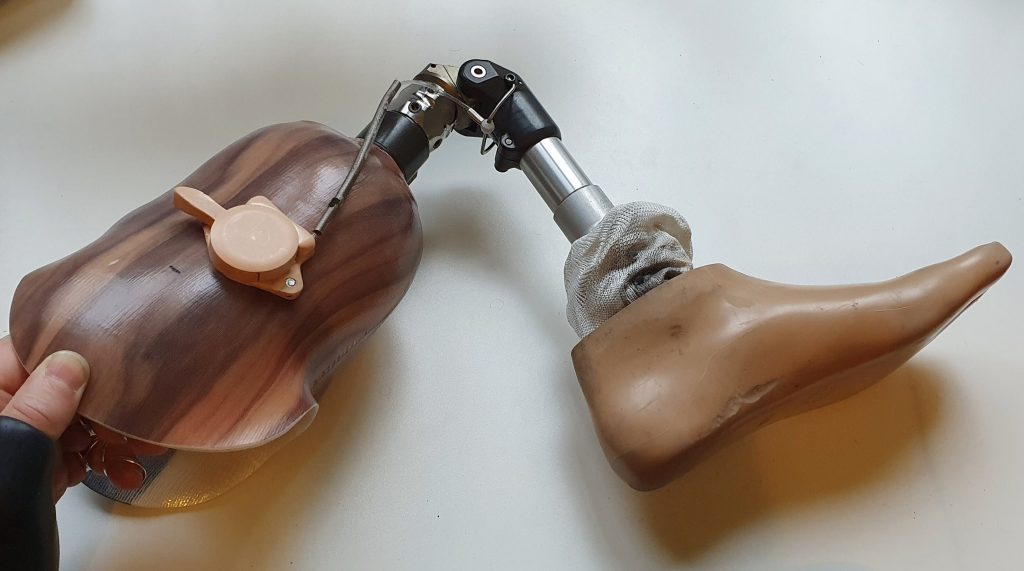 Afbeelding van een linker onderbeenprothese met een extra scharnier als knie-onder-knie-constructie die te bedienen is met een knopje op de zijkant van een houtlook koker waar de stomp na amputatie in past.