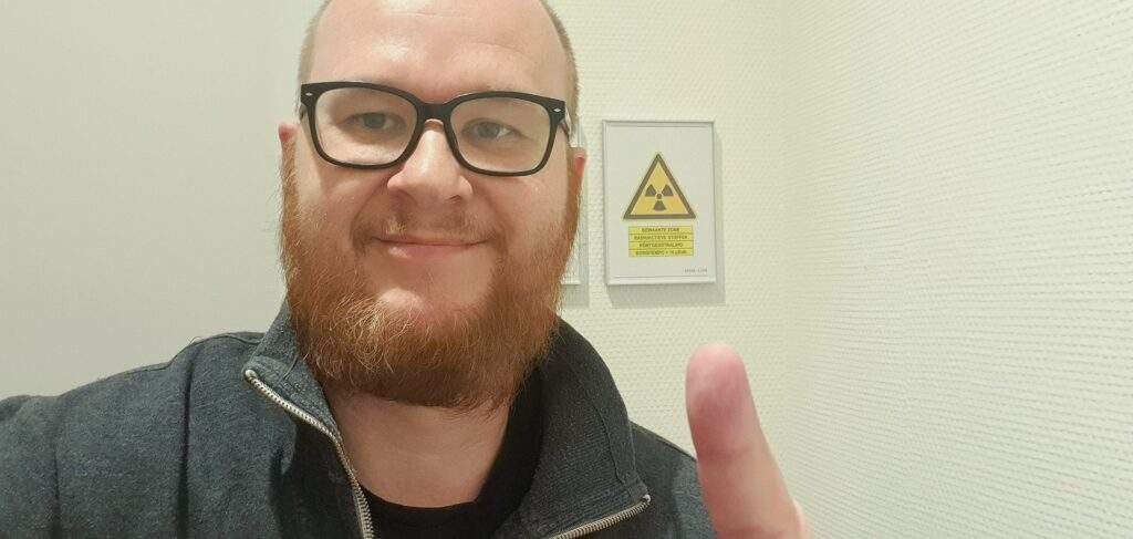 afbeelding van een man met baard bij een bordje waarop gewaarschuwd wordt voor een behandelruimte met nuceaire, radioactieve stoffen en röntgen straling, zoals bij een mag3 nierscan