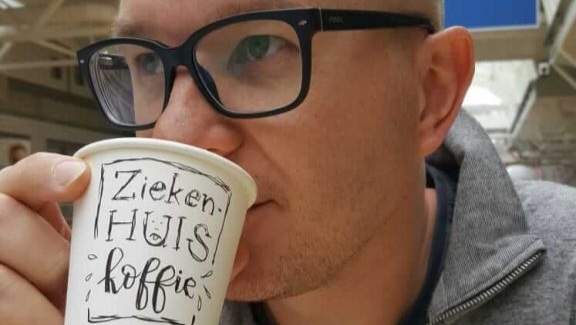 afbeelding van een man met bril die uit een wegwerpbekertje koffie drinkt met daarop met handlettering aangebracht de tekst 'Ziekenhuiskoffie'