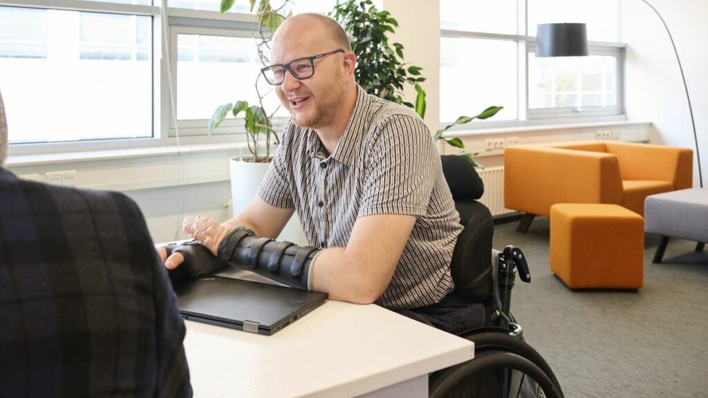 Afbeelding van Ralph Stoove die in zijn rolstoel aan een vergadertafel zit te overleggen. Voor hem staat een dichtgeklapte laptop. Het overleg werd gehouden voor Diversity Week waarbij er aandacht was voor werken met een beperking.