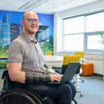 Foto van Ralph Stoové in een rolstoel met een laptop op schoot. Ralph zit in een kleurrijke kamer van een kantoor. Hij draagt een overhemd en een spijkerbroek en spalken om zijn handen en vingers.