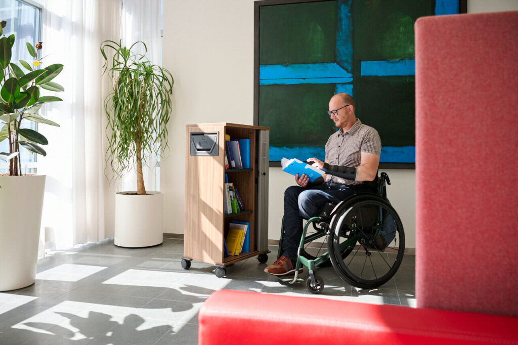 Ralph Stoove leest in een boek naast een verrijdbaar boekenkastje in de toegankelijke bibliotheek van een kantoor. Ralph zit in een rolstoel. Ralph heeft een amputatie van zijn linkerbeen en je ziet ook zijn spalken aan zijn handen en vingers, evenals de orthopedische schoen aan zijn rechter voet. Zijn groene actieve handbewogen rolstoel is een TNS Activator 2.5.
