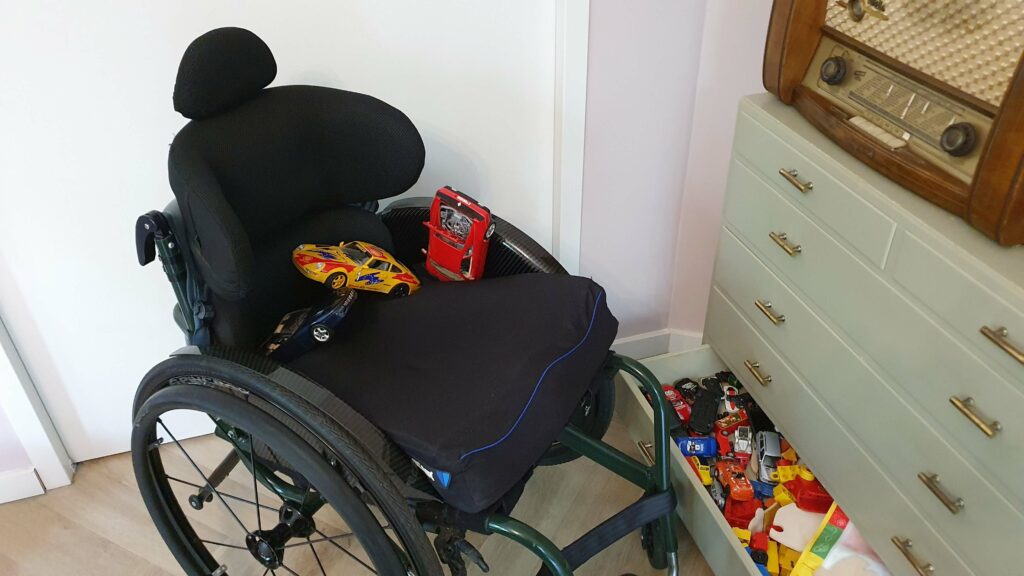 Afbeelding van een rolstoel zonder een persoon er in. Wel zie je drie grote speelgoedautootjes als gevonden voorwerpen in een rolstoel zitten. De rolstoel staat naast een dressoir waarvan een lade open is met allerlei speelgoedautootjes.