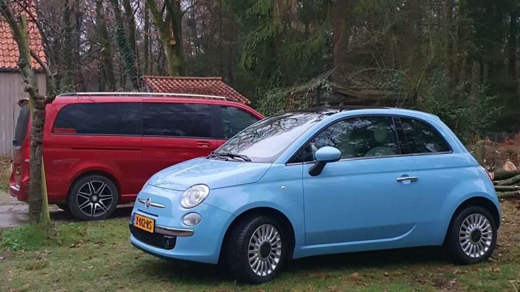 Foto van een Nuova Fiat 500 die geparkeerd staat in een bosrijke omgeving. Naast het lichtblauwe Fiatje staat een rode Mercedes-Benz V-Klasse geparkeerd. Zowel qua kleur als formaat contrasteert het kleine boodschappenautootje enorm met de grote rolstoelbus.