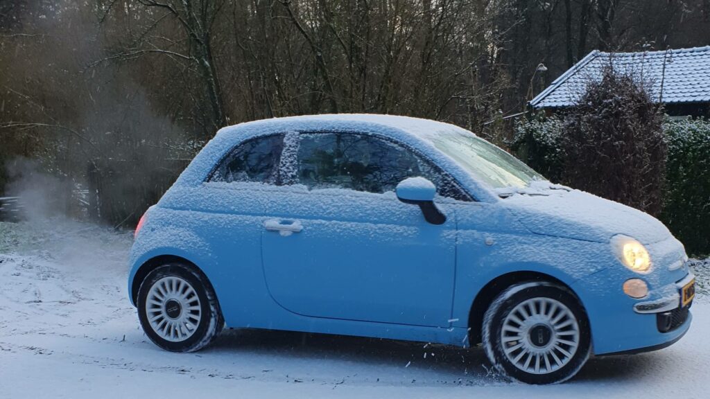 Foto van een accelererende, lichtblauw Fiatje in de sneeuw. je ziet aan de voorwielen de beweging en er spat sneeuw omhoog. De bijnaam van de auto is 'Bolletje'.