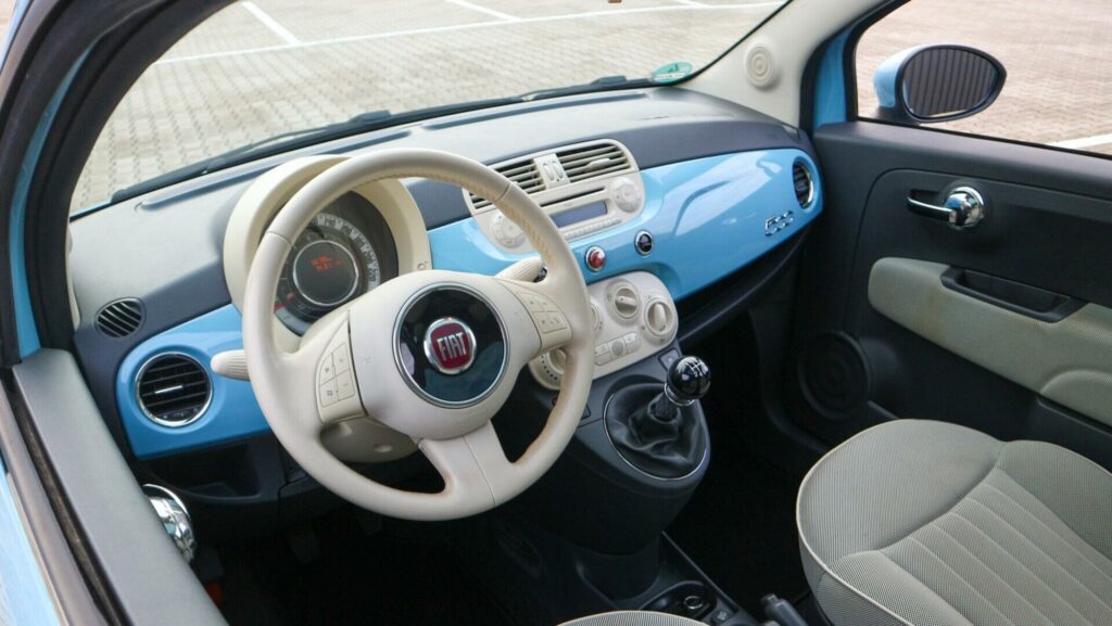 Afbeelding van het mooie, lichte en vrolijke interieur van een Nuova Fiat 500 Lounge uit 2013. Aan de kleur van het dashboard kan je zien dat dezelfde lichtblauwe kleur ook de kleur is van de auto. Het leren stuur, enkele panelen en knoppen zijn in beige wit uitgevoerd.