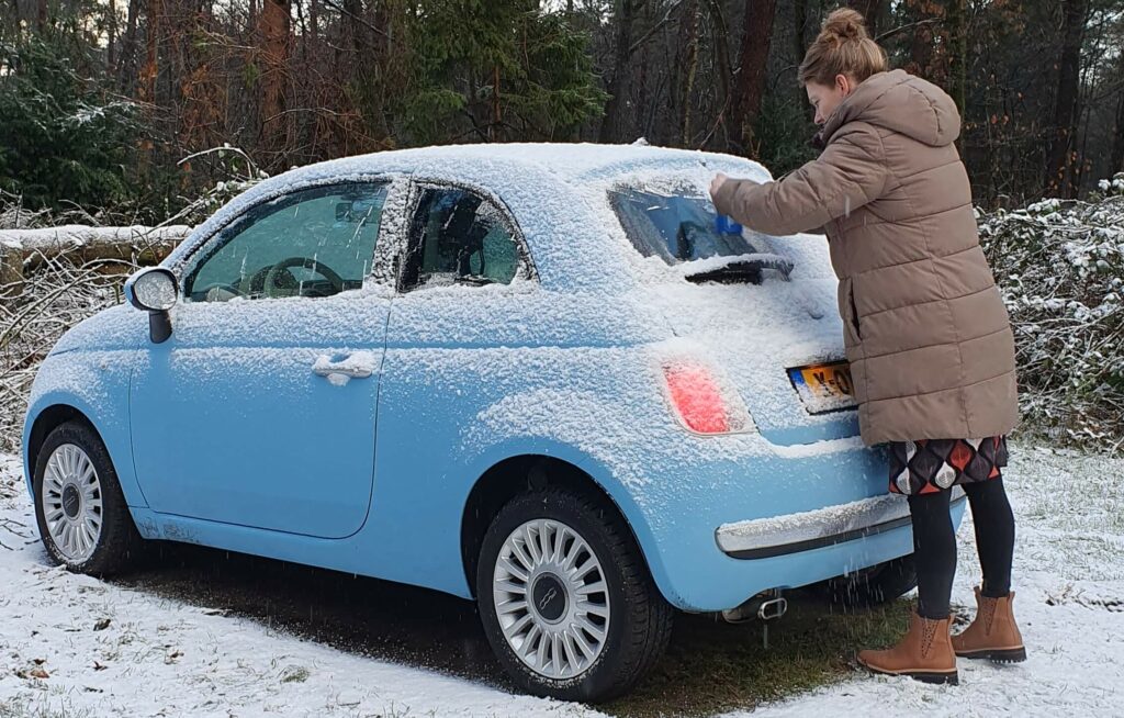 Afbeelding van een Nuova Fiat 500 uit 2013 in de babyblauwe kleur 'Legends Blue' en lichtmetalen velgen. Een mooie vrouw is de ruiten aan het krabben die vol met sneeuw en ijs zitten, om daarna veilig de weg op te kunnen.