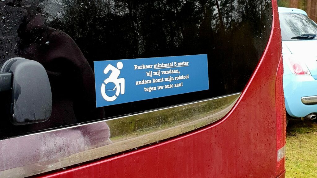 Foto van een rolstoelsticker op de achterruit van een rode bus met de grappige tekst 'Parkeer minimaal 3 meter bij mij vandaan, anders komt mijn rolstoel tegen uw auto aan!'