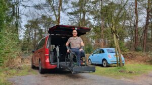 Foto van Ralph Stoove op de plateaulift van zijn rode Mercedes-Benz V-Klasse met de achterklep omhoog. Uit de achterkant komt een rolstoellift om hem in een rolstoel naar binnen te helpen, zodat diegene met een beperking of handicap kan autorijden. Het plateauliftje staat iets omhoog op dezelfde hoogte als de laadvloer van de rolstoelbus. Op de achtergrond zie je nog een modernde lichtblauwe Fiat 500. De omgeving is bosachtig.