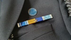 Close-up van een uniformjasje met daarop drie batons die aan elkaar zijn opgemaakt. Van links naar rechts zie je deze drie onderscheidingen: Links een donkerblauw met witte baton voor 12,5 jaar trouwe dienst, in het midden een geel met blauwe baton waarop een zilver ster in het midden is geplaatst voor moed en doorzettingsvermogen tijdens een uitzending voor het ministerie van Veiligheid en Justitie en rechts een lichtblauw met witte baton van de Verenigde Naties die ook wel United Nations Special Service Medal wordt genoemd. In het midden boven de drie batons zit een herinneringsspeld van een specialistische eenheid.