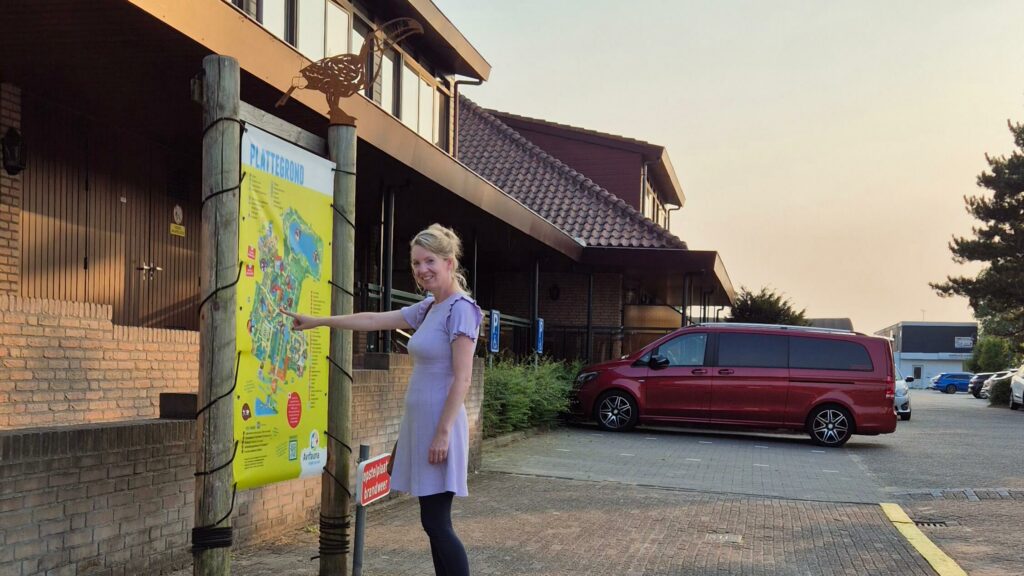 Afbeelding van een een vrouw bij het informatiebord van vogelpark Avifauna met op de achtergrond het mooiste rolstoelbusje van Nederland dat geparkeerd staat in één van de vele en ruime gehandicaptenparkeerplaatsen van Van der Valk Hotel Avifauna. De vrouw is blond en draagt een lila jurkje, de rolstoelbus is rood en van het merk en type Mercedes-Benz V-Klasse.