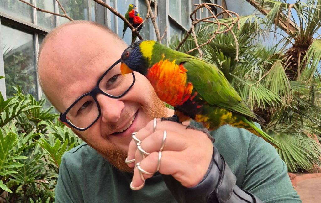 Een papegaai op de hand van Ralph. Ralph draagt een polshandspalk en silversplints om zij  vingers en heeft een bril op. De papegaai is felgekleurd met groen, oranje, geel en blauw, De foto is gemaakt in vogelpark Avifauna.