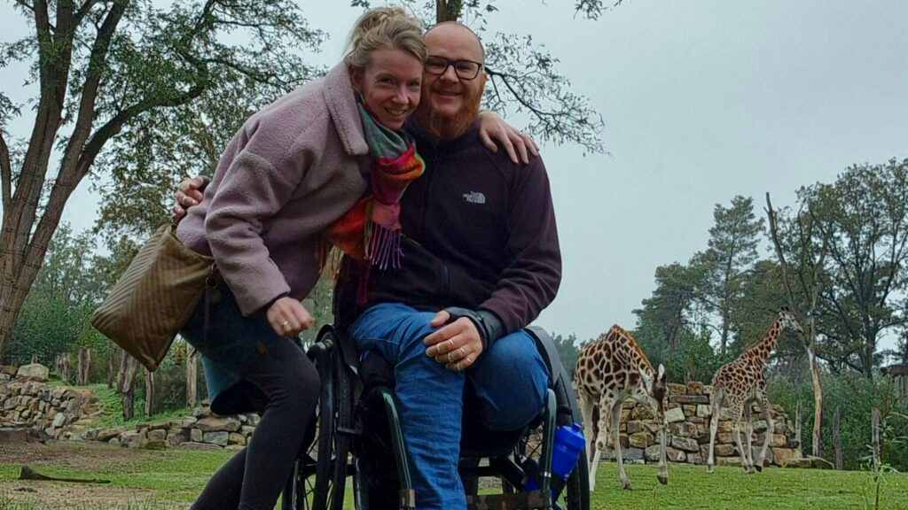 foto van Ralph Stoove in rolstoel met daarnaast zijn vrouw Marjolein in safaripark Beekse Bergen. Rechts op de achtergrond zie je giraffen grazen. Marjolein draagt een lila wollen jasje. Ralph draagt een zwart fleecejack en een blauwe spijkerbroek. De rolstoel van Ralph is groen.