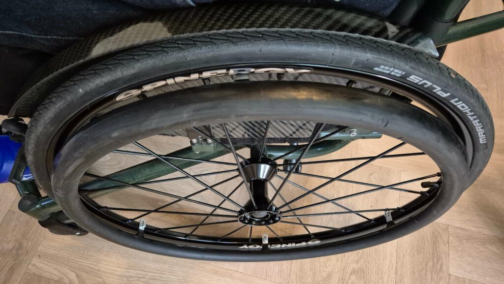 Een nieuw gemonteerd rolstoelwiel op een groene maatwerk rolstoel. Het wiel is van het merk Spinergy Wheels en de spaken, velg en naaf zijn zwart. De velg is 26 inch en afgemonteerd met een nieuwe Schwalbe Marathon Plus band. Er zit wel een gebruikte zwarte rubbergecouate hoepel op van het merk en type Carbolife Quadro.