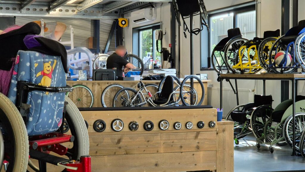 Afbeelding in de showroom van de hoofdvestiging van TNS Revalidatie Service in Dongen met in de achtergrond een monteur die bezig is met een rolstoelwiel, daarvoor op een sokkel een handbike met daaromheen maatwerk rolstoelen en op een houten wand verschillende soorten voorwieltjes voor een rolstoel.