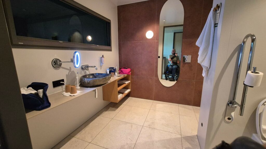 foto van de badkamer in de rolstoeltoegankelijke hotelkamer van Hotel Beekse Bergen. Links is een ruim en onderrijdbaar stenen aanrechtblad gemonteerd met daarop een wasbak waar je met de rolstoel onder kunt. Er is ook voldoende plek om je tandenborstel, verzorgingsproducten en medische spulletjes zoals stomamateriaal neer te leggen. Aan de wand hangt een passpiegel en bij de wasbak hangt een klein rond spiegeltje. In de reflectie van de grote spiegel aan de wand zie je Ralph Stoove een foto maken vanuit zijn rolstoel.