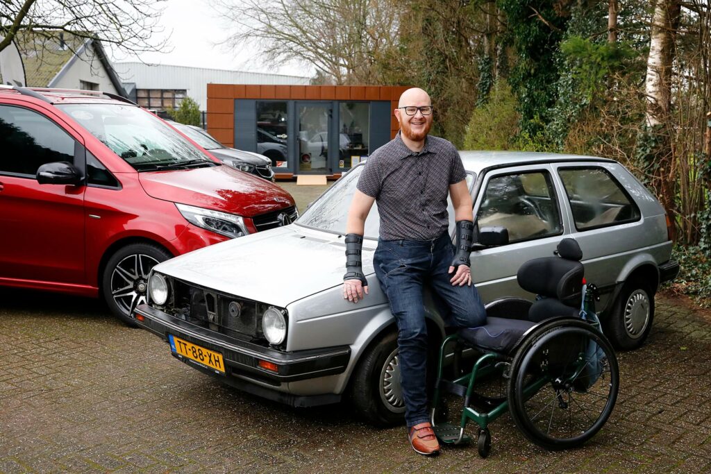 Foto van autoliefhebber Ralph Stoové leunend tegen zijn zilvergrijze VW Golf Mk2 oldtimer uit 1988 met op de achtergrond ook zichtbaar zijn rode Mercedes-Benz V-Klasse rolstoelbus. Op de voorgrond zie je ook de rolstoel van Ralph Staan. De rolstoel is dongergroen. Ralph draagt een bruin overhemd met ruitjesmotief, donkere spalken aan zijn armen, een donkerblauwe spijkerbroek waarbij zichtbaar is dat zijn linker been is geamputeerd én hij draagt een bruin met blauwe orthopedische schoen.