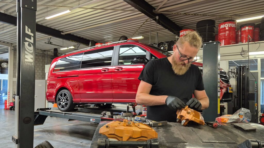 Foto van Maciek Kabza die bezig is met het klaarmaken van een W447 big brake kit voor op een Mercedes-Benz V-Klasse rolstoelbus. Die rolstoelbus is op de achtergrond te zien op een brug van de garage. Op de voorgrond ligt een remklauw van de remmenupgrade. Die is net zo goudkoper gespoten als AMG op de auto's toepast. Er zit nog geen AMG logo op de remklauwen.