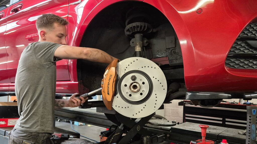 Foto van een monteur die een W447 big brake kit op de vooras van een Mercedes-Benz V-Klasse monteert. De remklauw is kopergoud en heeft de tekst en het logo van AMG erop. De remschijven zijn 375 millimeter en zijn geperforeerd. De bus zelf is rood.