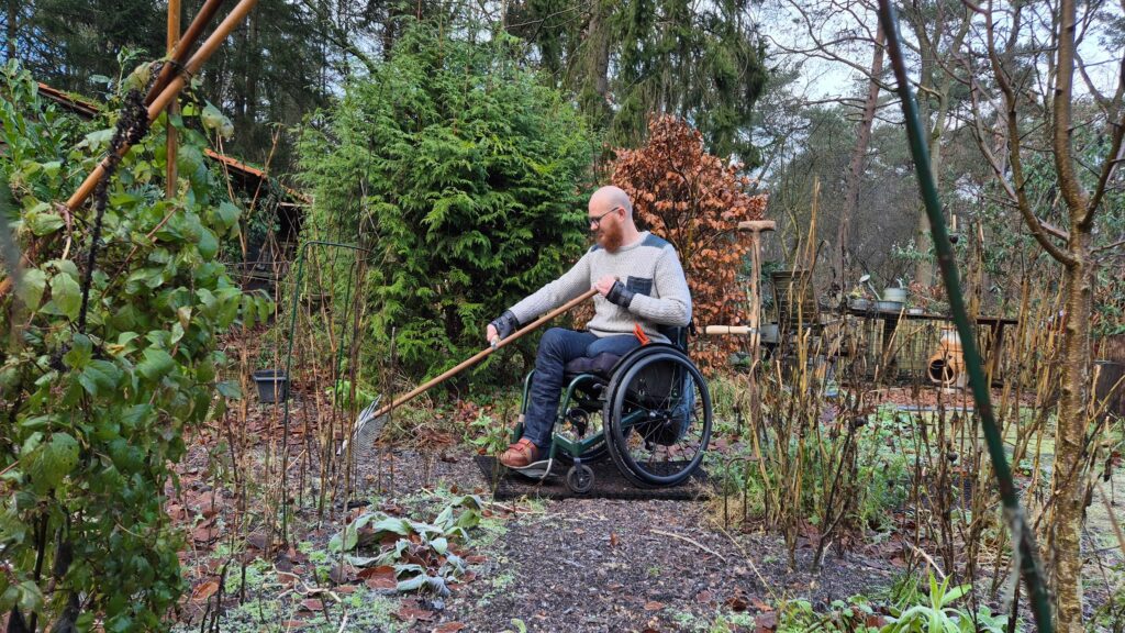Ralph Stoové zit in een rolstoel en harkt de tuin met aangepast tuingereedschap. De rolstoel staat op rubber gatenmatten. Ralph heeft links een bovenbeenamputatie en draagt om zijn handen en vingers spalken en silversplints. De foto toont een herfstbeeld van een natuurlijke tuin.
