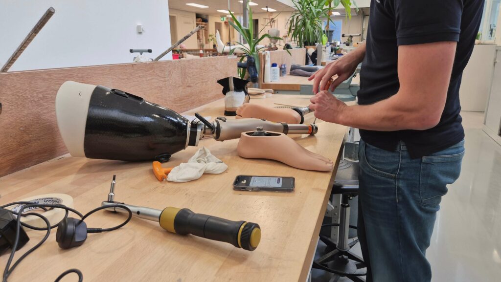 Een prothesemaker van ForMotion Orthopedie monteert een computergestuurde Kenevo knie met microprocessor op een beenprothese met een direct socket stompkoker. Op de werkbank ligt de prothese helemaal gedemonteerd waarbij je de koker, de protheseknie en de losse voeten ziet liggen met daaromheen gereedschap.