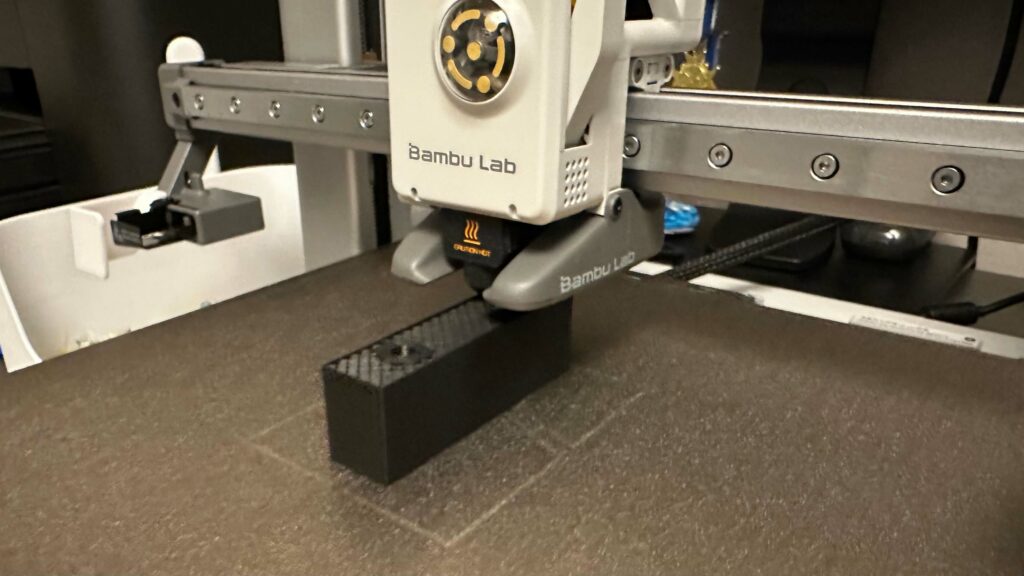 Afbeelding van een Bambu Lab 3D-printer die bezig is met het printen van een zwart blokje met twee gaten. Dat blokje moet uiteindelijk een voetsteuntje worden voor op een elektrische rolstoel van merk en type Quickie Q700M