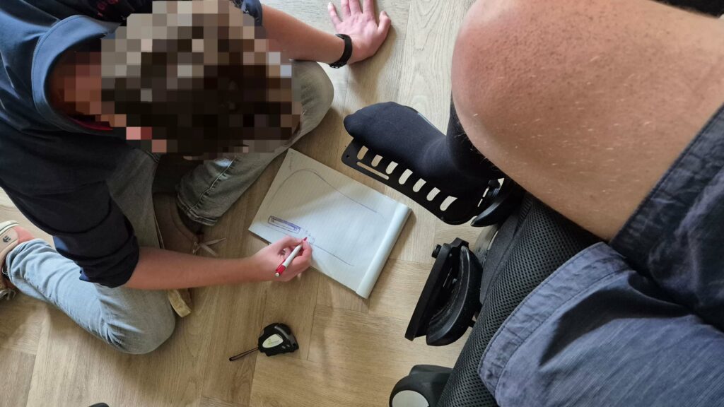 Foto van een geanonimiseerde adviseur van Medipoint die de maten oneemt en een ontwerpschets maakt voor het vervaardigen van een voetsteuntje op een elektrische rolstoel. Je ziet links de medewerker van Medipoint op de grond zitten met een kladblok met notities en een schets en een meetlint. Rechts zie je het verlamde rechterbeen van een man in korte broek die in een elektrische rolstoel zit. De voet rust op de voetplaat van de elektrische rolstoel. Links heeft de man ivm een bovenbeenamputatie geen been. De visgraat vloer is houtkleurig.