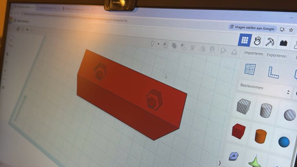 Afbeelding van een computer beeldscherm met daarop het ontwerp voor een 3D-printer van een blokje in een rode kleur met twee gaatjes en bovenaan die gaatjes twee zeskantige uitsparingen om twee boutjes te fixeren. Dat blokje is het ontwerp van een voetsteuntje voor op een elektrische rolstoel.