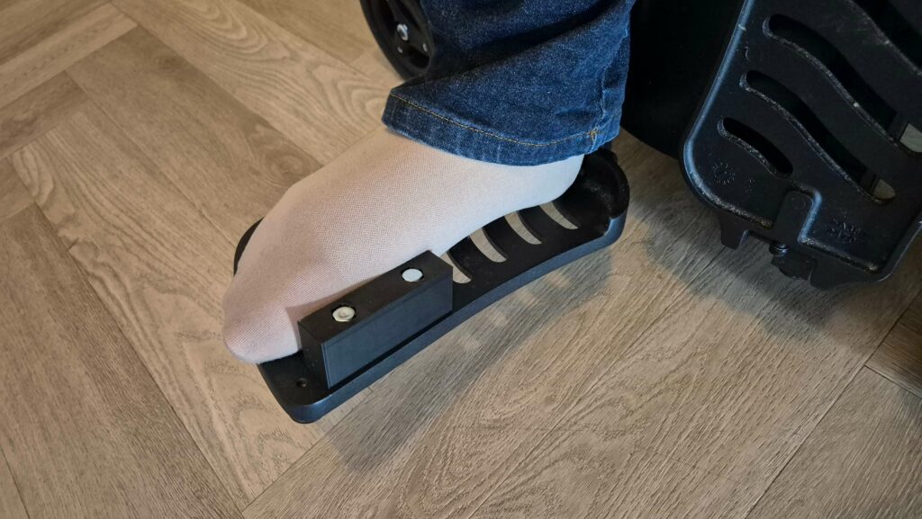 Afbeelding van een voetplaat van een elektrische rolstoel van het merk en type Quickie Q700. Op de voetplaat rust een verlamde voet in een vleeskleurige steunkous. Op de voerplaat zit een voetsteuntje waartegen de binnenzijde van de voorvoet rust.