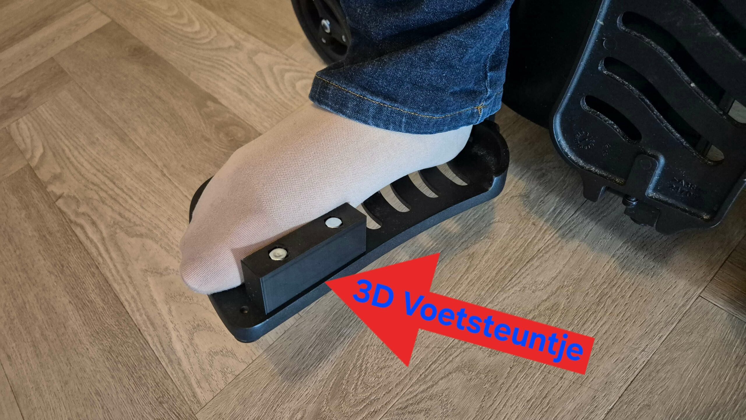 Afbeelding van een voetplaat van een elektrische rolstoel van het merk en type Quickie Q700. Op de voetplaat rust een verlamde voet in een vleeskleurige steunkous. Op de voerplaat zit een voetsteuntje waartegen de binnenzijde van de voorvoet rust. Naar het voetsteuntje wijst een rode pijl met de blauwe tekst '3D Voetsteuntje'.