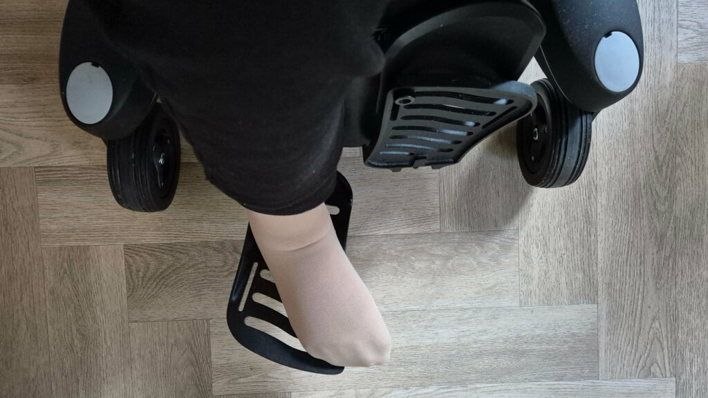 Afbeelding van een voet die verlamd is in een vleeskleurige steunkous. De voet rust op een voetplaat van een Quickie Q700 elektrische rolstoel, maar staat duidelijk scheef, omdat door de verlamming de voet naar binnen draait. De foto is van bovenaf genomen. De elektrische rolstoel is zwart en de visgraat vloer is houtkleurig.