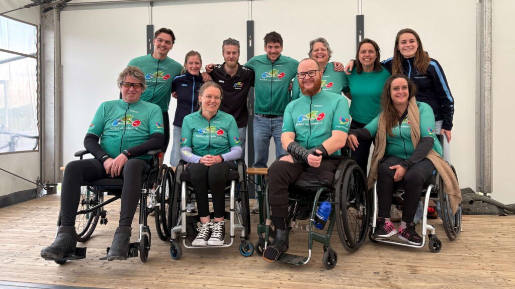 Afbeelding van het 2026 Birds on Wheels handbiketeam van revalidatiecentrum Vogellanden. Dit is de presentatie van de teamshirts. Die shirts zijn een beetje mintgroen met het teamlogo erop. Vooraan zie je ver rolstoelgebruikers, waarvan de derde van links Ralph Stoové is. Achter staan andere teamleden, buddy's en handbikers die niet rolstoelgebonden zijn.