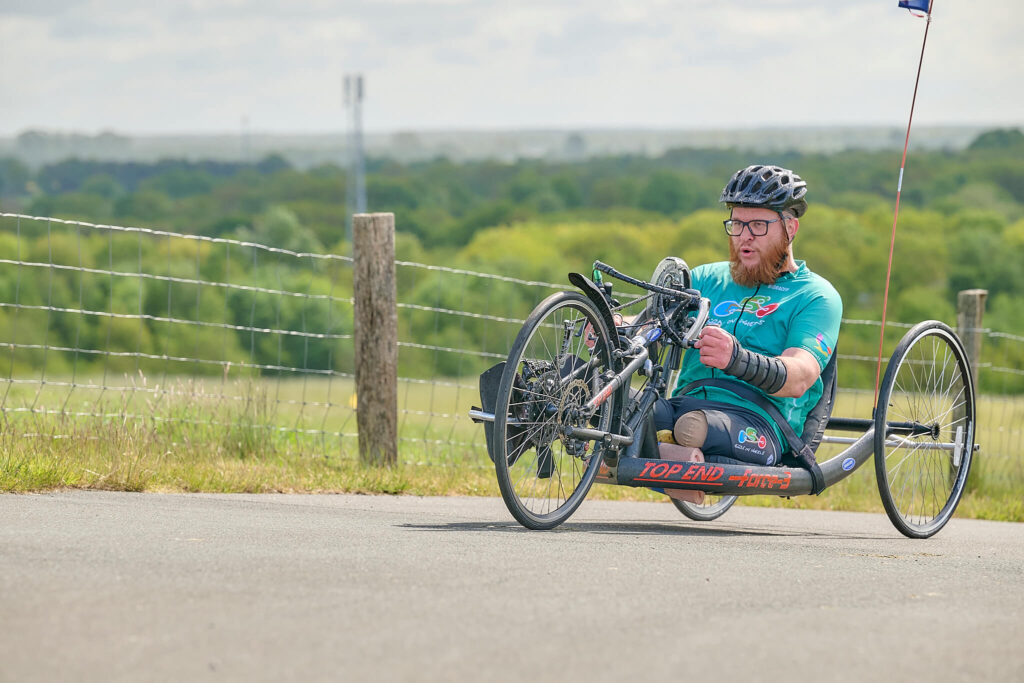 Afbeelding van Ralph Stoove die op een vastframe race handbike rijdt. Ralph heeft een teamshirt en broekje aan van Birds on Wheels Vogellanden met als hoofdkleur mintgroen/turguoise. Ralph kijkt heel geconcentreerd terwijl hij een heuvel op rijdt. Hij draagt ook een zwarte fietshelm en donkere siliconen polshandduimbraces. Zijn linkerbeen is hoog boven de knie geamputeerd. De handbike is zwart met rode letters met de tekst 'Top End Force 3'.