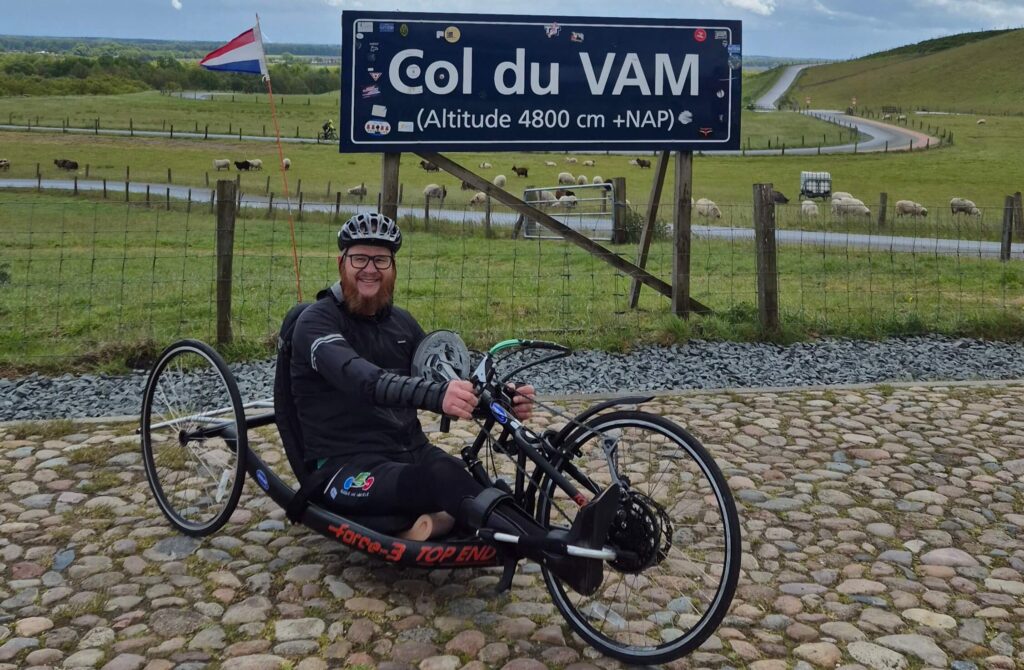 Afbeelding van een man (Ralph Stoové) in een vastframe race handbike bij het bordje van de Col du Vam met daarop de tekst 'Col du Vam 4800 cm +NAP'. Op de achtergrond zie je het glooiende landschap en weilanden. Ralph staat met zijn handbike op kasseien. De handbike is zwart met rode letters. Ralph draagt een zwarte outfit met op zijn fietsbroek het logo van zijn team zichtbaar, namelijk 'Birds on Wheels Vogellanden'. Ralph heeft rechts een verlamd been met een enkelvoetorthese en links heeft hij een hoge bovenbeenamputatie.