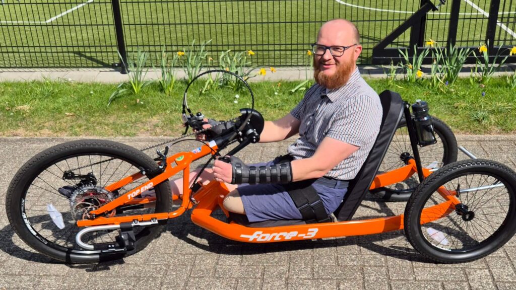 Een man met links een geamputeerd been zit met een korte broek en een overhemd in een oranje vastframe handbike van merk en type: Top End Force 3.