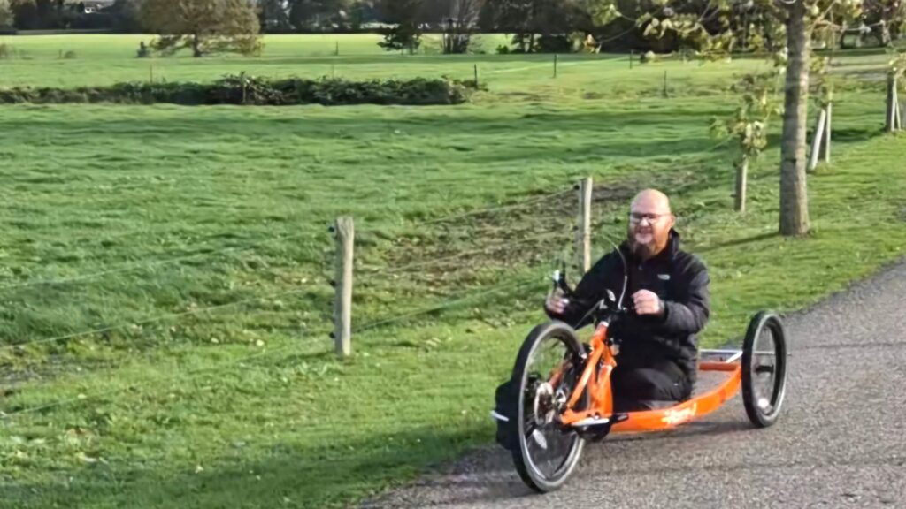 Screenshot van een video waarop een man, Ralph Stoové, te zien is die rijdt met een oranje vastframe handbike. Op de achtergrond zie je een landelijke omgeving. De man heeft links een hoge bovenbeenamputatie, rechts een deels verlamd been in een spalk en ook om zijn handen draagt hij spalken. De man is kaal, maar heeft ook een baard. Hij draagt een donkere broek en een zwarte jas.