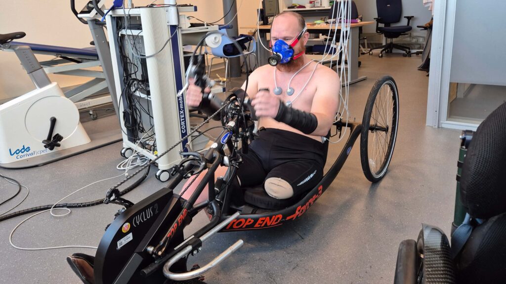 Foto van Ralph Stoové tijdens de sportmedische keuring voor de Handbike Battle 2026. De zwart met rode Top End Force 3 vastframe handbike is geprepareerd in een statische opstelling voor het meten van de conditie. Ralph heeft een ontbloot bovenlijf met daarop allerlei draden om zijn vitale functies te monitoren. Ook draagt Ralph een masker op zijn gezicht om de samenstelling van zijn adem te onderzoeken tijdens inspanning. Ralph heeft een strakke sportbroek aan en heeft links een hoge beenamputatie. Ook draagt Ralph spalken om zijn handen en polsen.