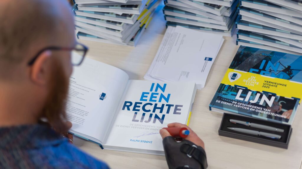 Close-up van auteur Ralph Stoové die het geschiedenisboek 'In een rechte lijn vernieuwde uitgave 2025' signeert. De foto is gemaakt over de rechter schouder van Ralph. Ralph heeft een kaal hoofd en een rode baard. Voor hem ligt het boek opengeslagen waarbij de titel 'In een rechte lijn' duidelijk te lezen is met daaronder een handtekening. Ralph heeft een pen vast met een verdikt handvat ivm zijn handbeperking. Op de achtergrond zie je meerdere stapels van dezelfde boeken klaarliggen voor verzending naar collega's en ketenpartners.