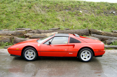 Foto van de zijkant van een Ferrari 328 GTS in de kleur Rosso Corsa. Op de achtergrond is een groene heuvel te zien en het asfalt is nat.