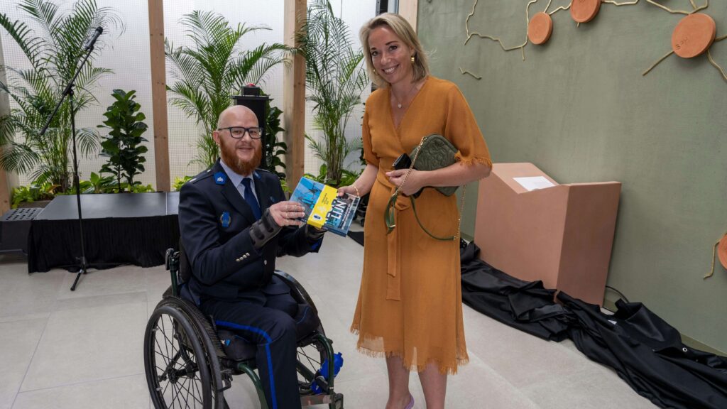 Afbeelding van de overhandiging van het boek 'In een rechte lijn, de geschiedenis van de Dienst Vervoer en Ondersteuning - vernieuwde uitgave 2025' door auteur Ralph Stoové aan voormalig staatssecretaris Ingrid Coenradie. Ralph draagt als uniform het ceremonieel tenue en zit in een rolstoel. Ralph draagt spalken en hij heeft links een hoge bovenbeenamputatie. Ingrid Coenradie draagt een mosterdgele jurk met een handtas.